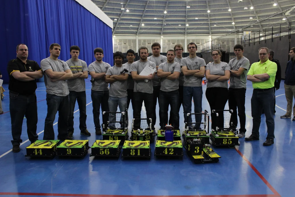 A group of people stands behind five numbered robots in a large indoor gymnasium, ready for an exciting Robot Football competition.