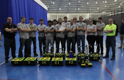 A group of people in gray shirts stand behind numbered robots on a blue indoor court, ready for the Valpo Football Combine.