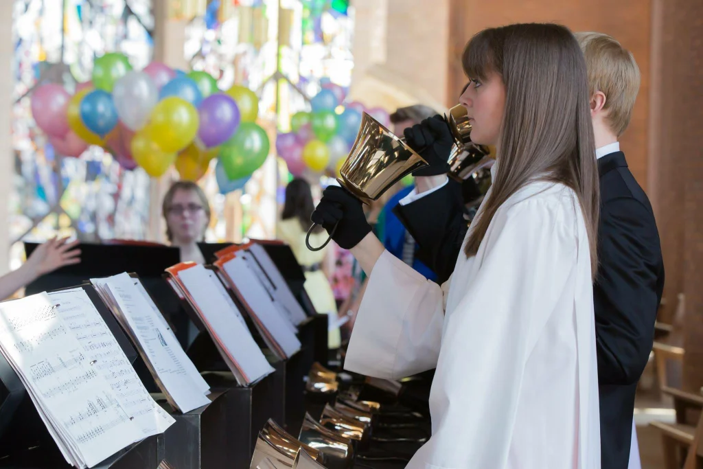 Choir member Kaitlin Werth plays a handbell in a church, with colorful balloons brightening the background.