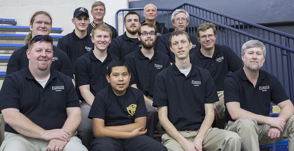 Group of thirteen men and one boy in matching black shirts, seated on bleachers and smiling at the camera during a Robot Football tournament.