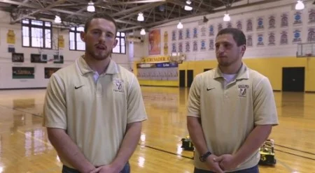 Two men in matching polo shirts stand in a gymnasium with Valpo banners and equipment in the background.