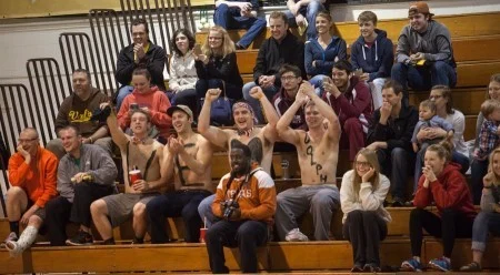 A group of excited fans, some shirtless, cheer in bleachers at an indoor Robotics Football event.