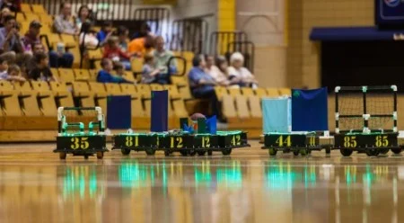 Small numbered carts with fabric panels on a gym floor; spectators sit in the bleachers as the Valpo Robotics Football Team prepares for their next match.