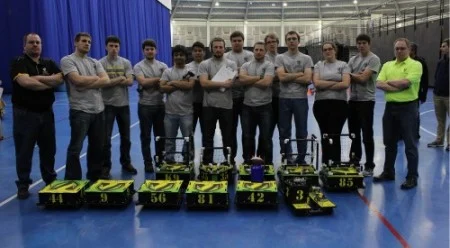 A group of people stands behind small yellow and black robots on a blue indoor court, ready for an exciting match of Robotics Football with the Valpo Robotics team.