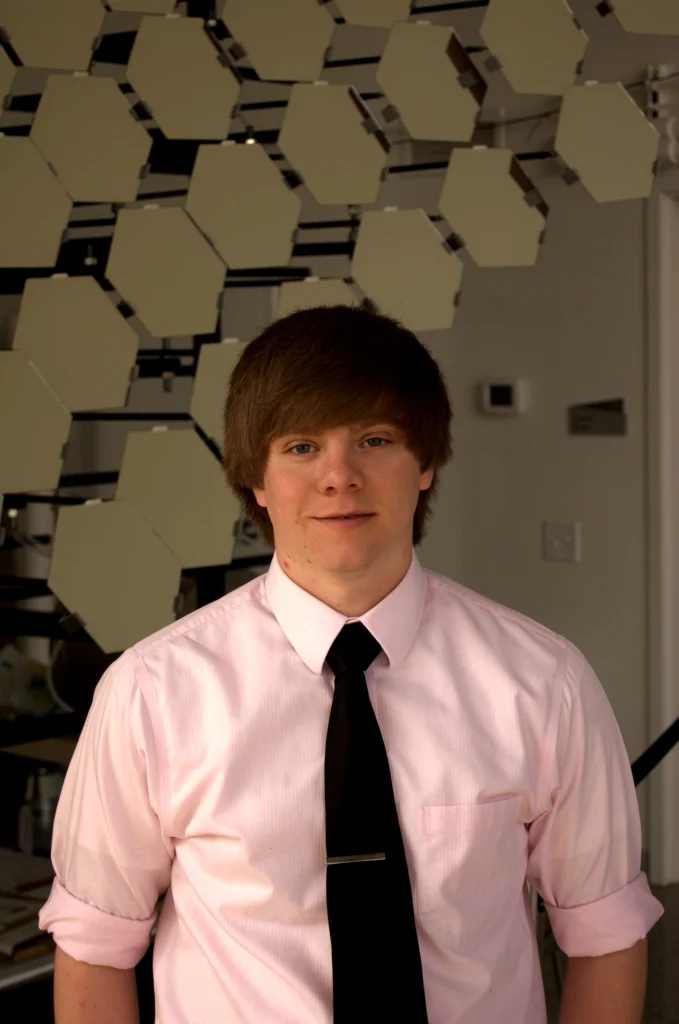 A young person in a pink shirt and black tie stands indoors in front of a geometric wall display.