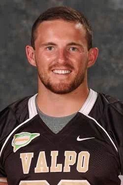 A smiling man in a black Valpo football jersey poses for a portrait against a gray background.