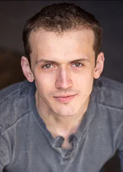 Young man with short brown hair in a grey shirt, looking up at the camera with a neutral expression—capturing a moment of life balance between profession and personal reflection.