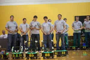 Students in gray shirts follow competition rules as they control numbered robots at a gymnasium event.