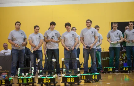 Students in gray shirts follow competition rules as they control numbered robots at a gymnasium event.