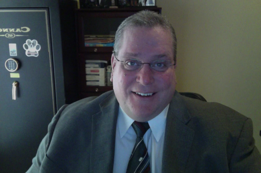 Smiling man in a suit and tie, possibly Thomas Biedron, sits in an office with shelves and a safe in the background.