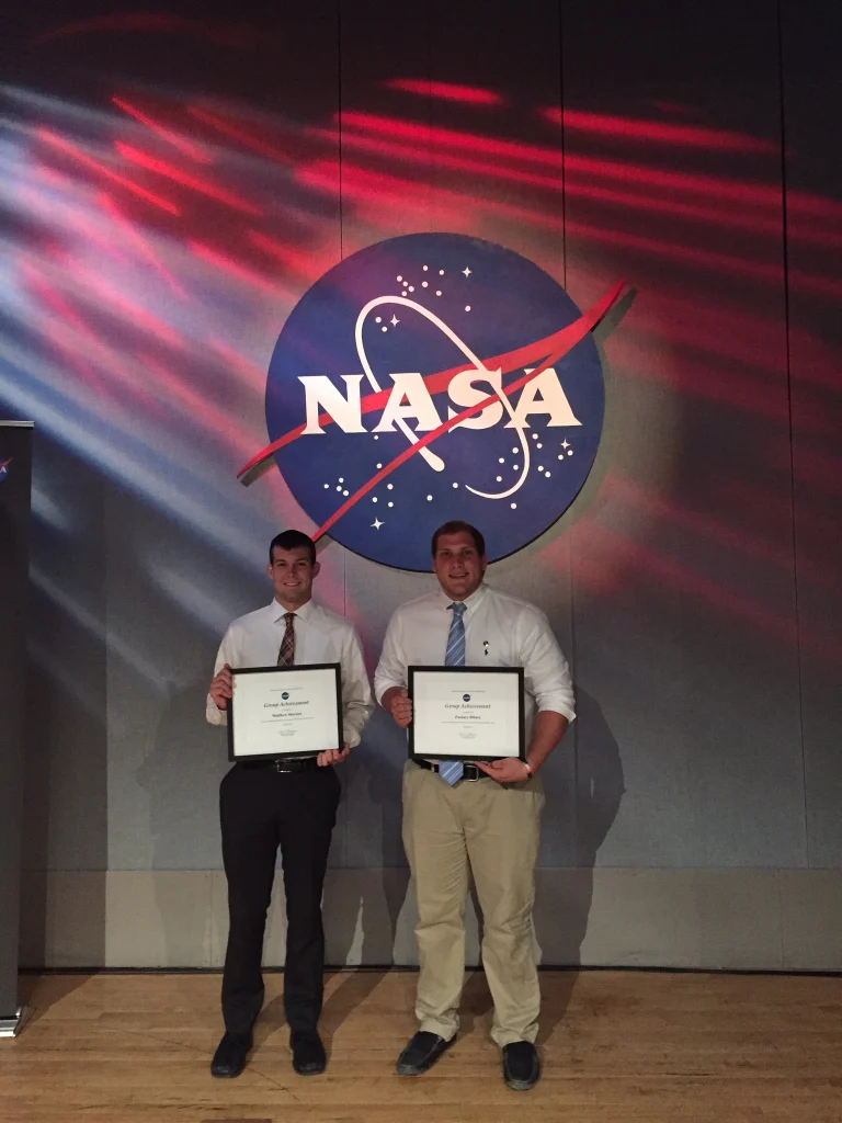 Two Valpo student interns stand holding certificates in front of a large NASA logo on a wall with red and purple lighting.