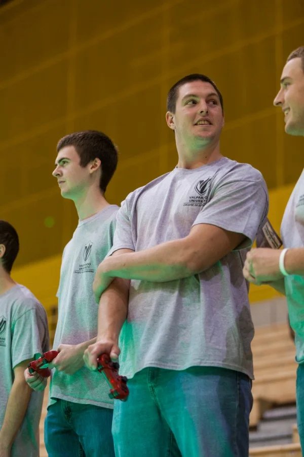 Three young men in matching gray shirts stand indoors, two holding video game controllers, chatting about ROBOT FOOTBALL and their recent match against Notre Dame.