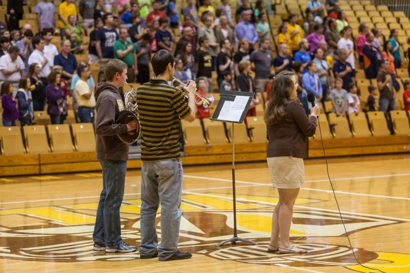 Three students from Valpo perform with instruments and a microphone in a gym during the Notre Dame ROBOT FOOTBALL event as a crowd stands and watches.