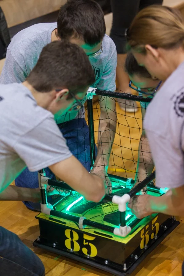 Three people work on a glowing green ROBOT FOOTBALL device with netting and the number 85 on its sides, representing the rivalry between Notre Dame and Valpo.