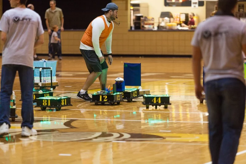 A person in sports gear oversees small numbered robots playing ROBOT FOOTBALL on a gym floor during an indoor Notre Dame event or competition.