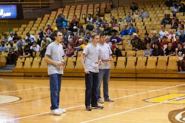 Three young men in gray shirts stand on a basketball court facing an audience in bleachers, as Valpo and Notre Dame fans gather for an exciting ROBOT FOOTBALL demonstration.