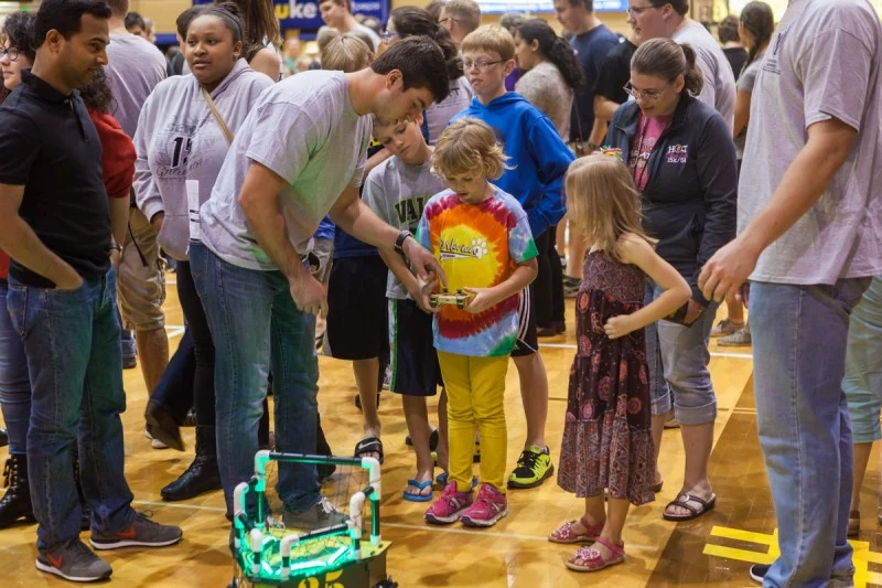 A group of kids and adults watch a boy operate a small robot at an indoor event, inspired by Notre Dame's ROBOT FOOTBALL excitement.