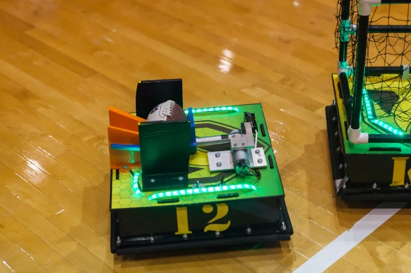 A small ROBOT FOOTBALL player, marked with the number 12 and glowing green lights, holds a football on the gymnasium floor—ready to represent Valpo in the match.