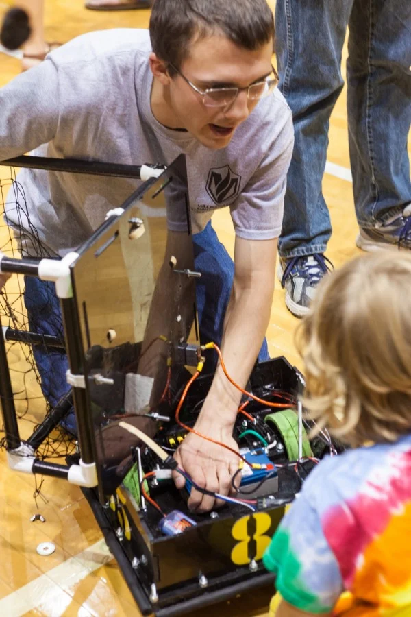 A person kneels on a gym floor, showing a child the inside of a small, open robot with wires and parts—an exciting moment from a Notre Dame vs. Valpo ROBOT FOOTBALL event.
