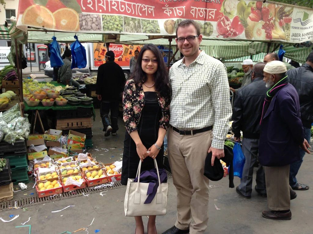 A man and woman pose in front of a fruit and vegetable market stall on a busy street, capturing the spirit of welcoming new colleagues in a vibrant community setting.