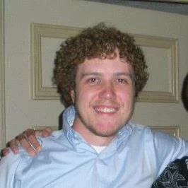 A smiling young man with curly hair wearing a light blue shirt stands in front of a beige door, looking as if he's ready to share stories from movie history or discuss classics like The Butler.