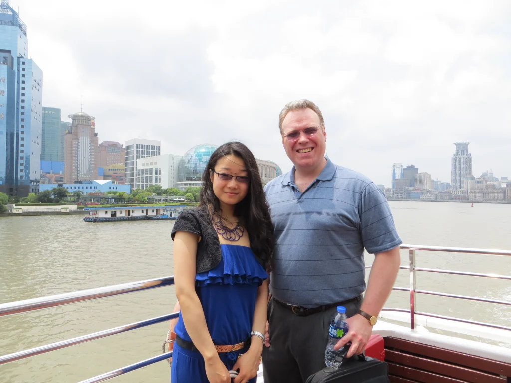 A man and woman stand on a riverside boat in Shanghai, with the city skyline behind them on a cloudy day—capturing a moment of making history together.