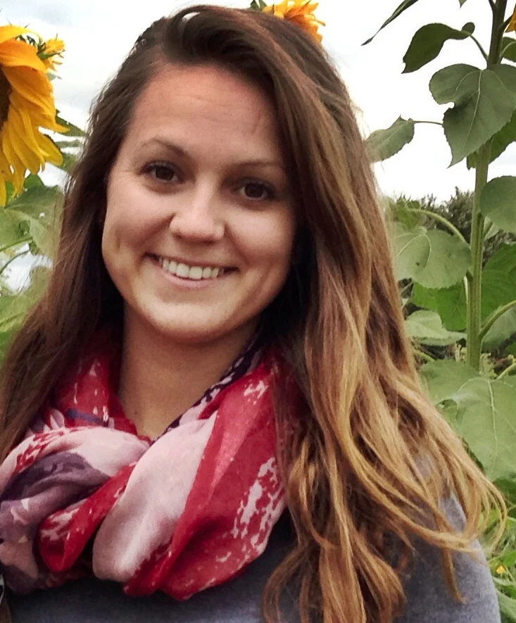 Smiling woman with long brown hair and a red scarf stands in front of sunflowers outdoors, reminiscent of a scene from History Goes to the Movies.