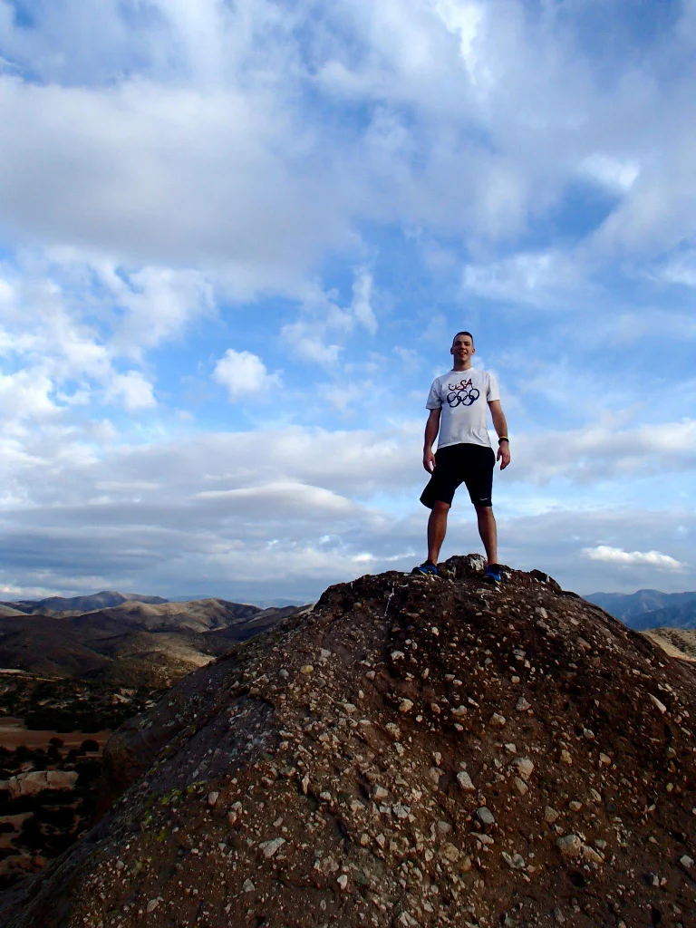 Man standing on a rocky hilltop under a blue sky with scattered clouds, overlooking a mountainous landscape, as if contemplating history like scenes from movies such as 12 Years a Slave.
