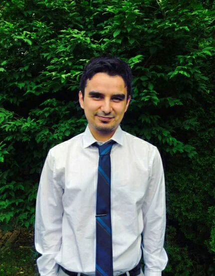 Professor J. Luis Ramos, Ph.D., in a white shirt and blue tie, stands smiling in front of green leafy bushes.