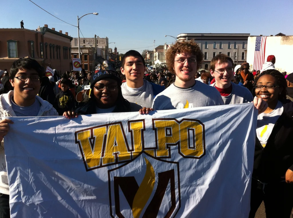 Six smiling students hold a large "VALPO" banner at an outdoor event, commemorating Civil Rights with a crowd gathered in the background.