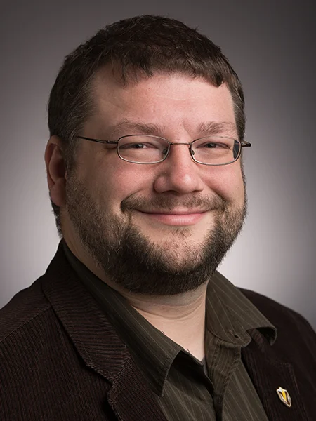 Alex Capaldi, a man with glasses and a beard, smiles at the camera, wearing a dark jacket and striped shirt. Known for his mentoring skills, he recently received a national award.