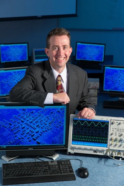 Man in a suit smiling, surrounded by monitors showing circuit boards and an oscilloscope displaying waveforms—highlighting Valpo Engineering's innovative environment for students.