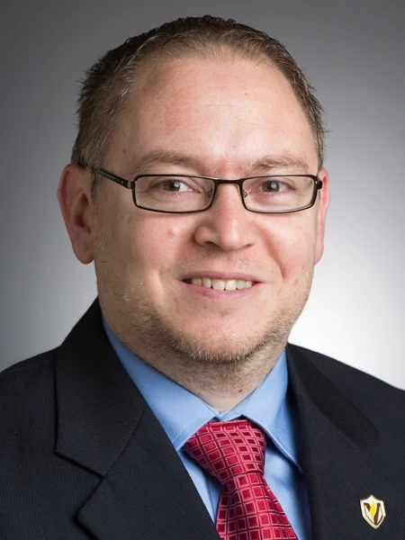 Man wearing glasses, a suit, blue shirt, and red patterned tie, smiling at the camera against a gray background; an expert in emerging technologies from Valparaiso University.