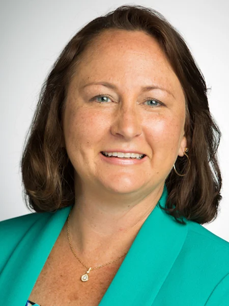 Smiling woman with shoulder-length brown hair wearing a teal blazer and a gold necklace, posing for a portrait, highlights her involvement in Wheat Ridge Ministries and dedication to expanding research opportunities.