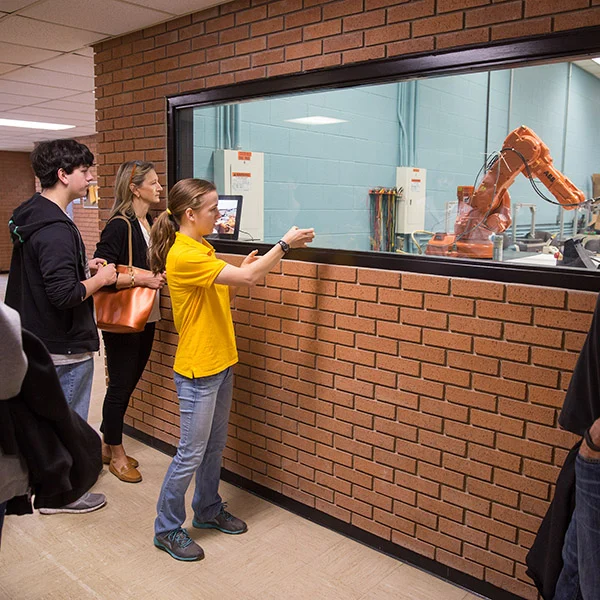 Engineering students observe an orange robotic arm working behind a glass window in a laboratory setting, highlighting hands-on experience that supports recruitment efforts.