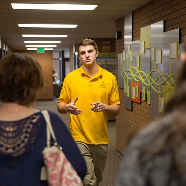 A young man in a yellow shirt discusses recruitment efforts with two people in a hallway adorned with art on the wall.