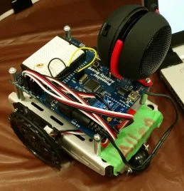 A small wheeled robot with a circuit board and a black-red speaker, designed for robotics research, sits next to a laptop.