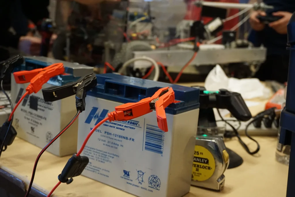 Close-up of batteries with jumper cables attached, surrounded by tools and equipment on a workbench at a Midwest Robotics Competition.