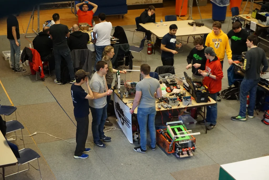 A group of people working on robotics projects at tables in a large indoor space prepares for the Midwest Robotics Competition.