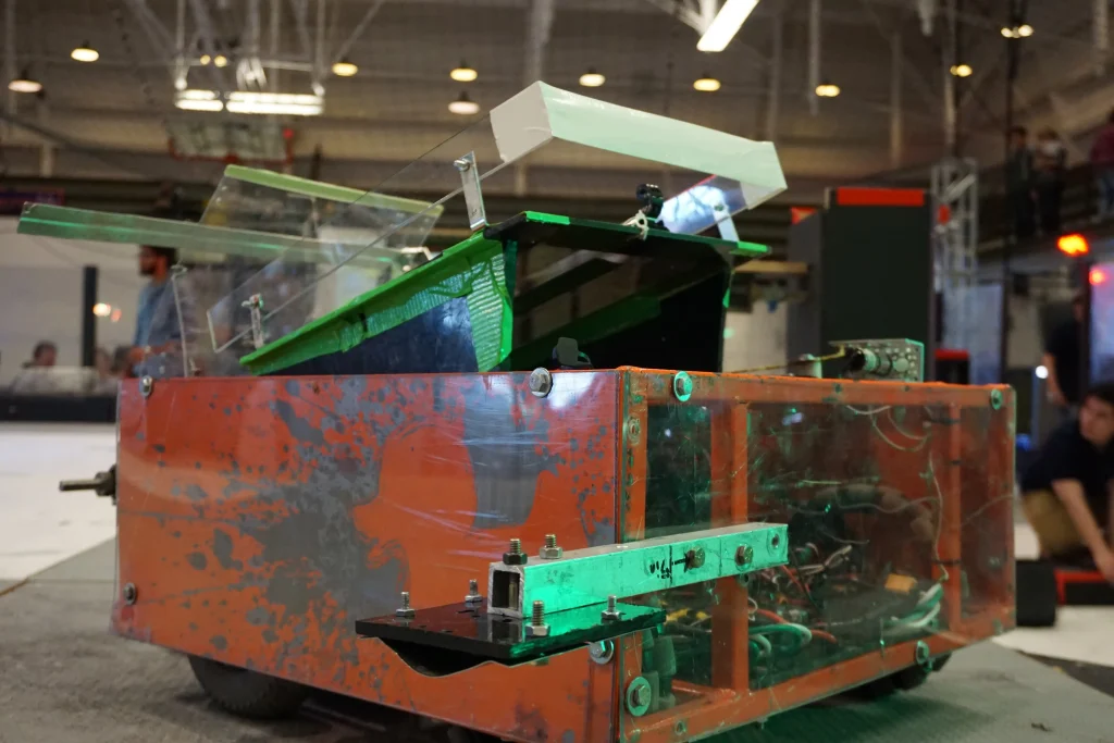 A red and green combat robot with a slanted metal top sits on an arena floor indoors, ready to showcase its Robotics Design skills at a Midwest Robotics competition.