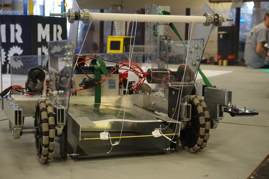 Close-up of a custom-built robot with mecanum wheels, wires, and transparent panels on a workshop floor—perfectly engineered for Midwest Robotics competition.