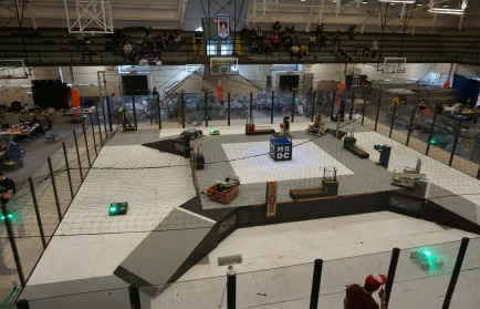 Robots compete inside a netted arena during the Midwest Robotics Competition, with spectators watching from the stands.