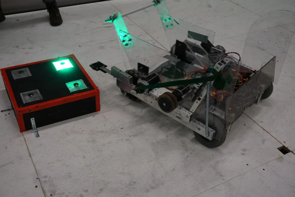 A small wheeled robot with green lights faces a black and red box with glowing green panels, showcasing innovative Robotics Design.