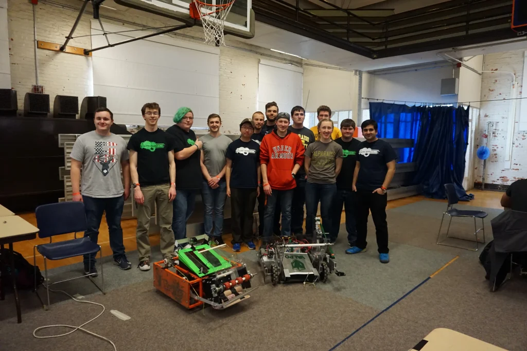 A group of people stand indoors behind two large robots on the floor, posing and smiling for a group photo at the Midwest Robotics Competition.
