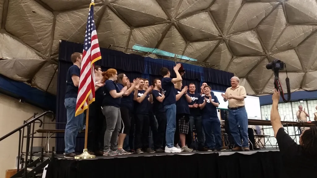 A group of people onstage clapping and smiling under a dome, with an American flag in the foreground during the Valpo Robotics Robotic Football Championship.