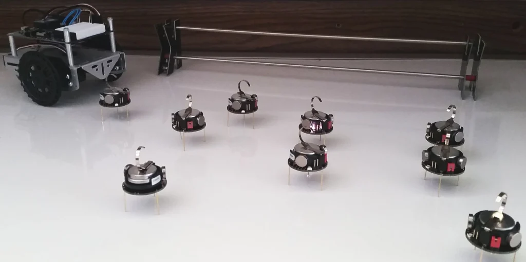 Seven small round robots with pins and hooks sit on a white surface near a larger wheeled robot, showcasing robotics research into swarm behavior.