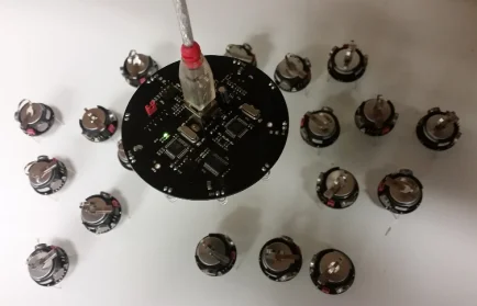 A round black circuit board with a USB cable, surrounded by small battery holders on a white surface—ideal for research in swarm robotics.