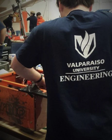 Person in a "Valparaiso University Engineering" shirt working on a project at a workshop or competition, representing Valpo Robotics.