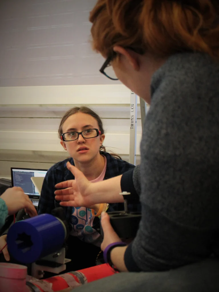 Two people from Valpo Robotics discuss a project with tools and machinery; one person listens intently, looking focused.