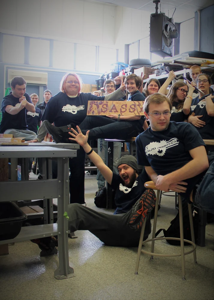 A group of people in matching shirts pose, one holding a large sign that reads "SASSY," smiling and cheering—proud members of Valpo Robotics celebrating their team spirit.